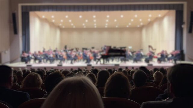 An out-of-focus orchestra on stage with silhouettes of the audience visible in the concert hall.
