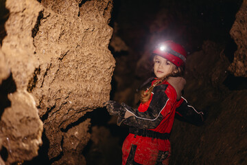 A 6-year-old girl in a protective suit and helmet with a flashlight in the middle of a natural underground cave is walking the tourist route of a caver