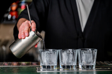 A bartender in a black jacket pours a dark liquid from a shaker into three glasses filled with ice. The setting is a vibrant bar with colorful bottles in the background.