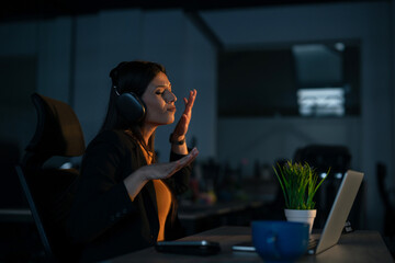 Happy Young woman in a modern office enjoying music through headphones while working late on a...