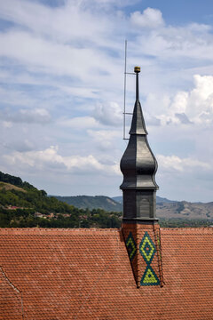 Stylized ventilation tower with lightning rod, installed on old red tiled roof of European town. Against background of bright blue expressive sky with fluffy clouds. Copy space for text.