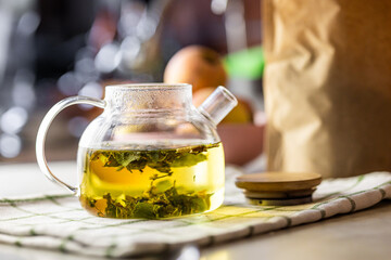 A clear glass teapot containing herbal tea with visible green leaves, resting on a checked cloth....