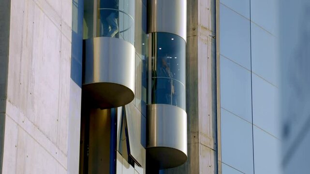 Futuristic glass elevators on the exterior of a modern high-rise building with a sleek, curved glass facade. The transparent design allows passengers to view the cityscape while ascending or descendin