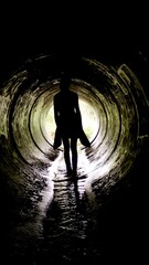A person walks through a dark drain tunnel, silhouetted against bright natural light at the tunnel's end, embodying the spirit of adventure and exploration in a unique setting
