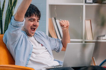 young man or student sitting indoors with laptop and euphoric expression of success