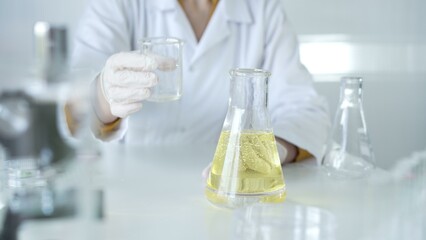 A scientist, wearing a lab coat and white protective gloves, is pouring a yellow oily liquid from one beaker to another in laboratory, close up. Medicine and science concept
