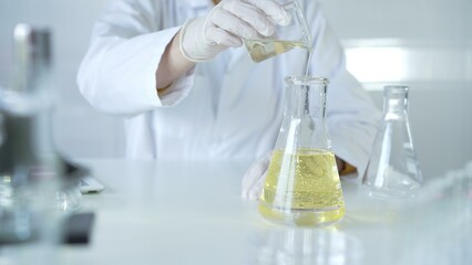 A scientist, wearing a lab coat and white protective gloves, is pouring a yellow oily liquid from one beaker to another in laboratory, close up. Medicine and science concept
