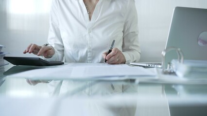 Businesswoman in blue blouse is using black calculator and taking notes while working on financial...