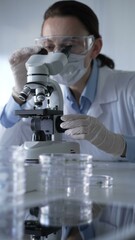 Woman scientist wearing white gloves, protective glasses and medical mask is using microscope doing research in modern laboratory setup. Low key lighting photo. Science and Medicine concepts