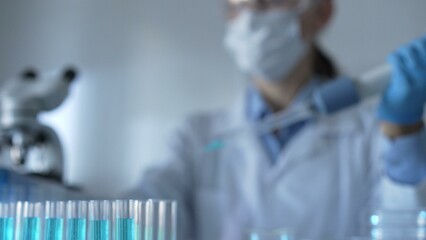 Test tubes with a blue liquid are in focus in laboratory setting, closeup view. Female scientist...