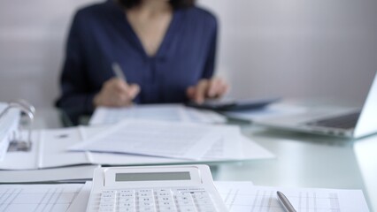 White calculator, pen and documents are lying on accountant's desk. Businesswoman wearing blue dress is working with documents on the background. Business and audit concept