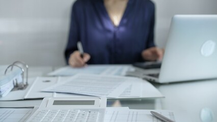 Magnifying glass, white calculator, pen and documents are lying on accountant's desk. Businesswoman wearing blue dress is working with documents on the background. Business and audit concept