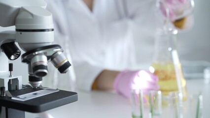 A scientist, wearing a lab coat and pink protective gloves, is pouring a yellow oily liquid from one beaker to another near microscope in laboratory, close up. Medicine and science