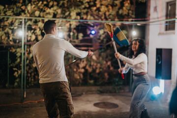 Two individuals engage in a playful broom sword fight outdoors, illuminated by festive string...