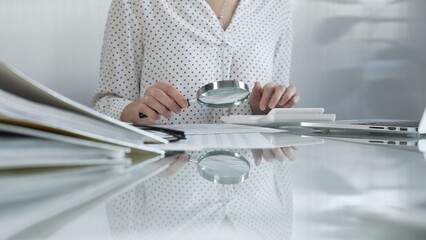 Businesswoman carefully reviewing financial reports with magnifying glass and calculator, conducting audit and analyzing data. Taxes, audit in business