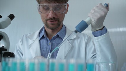 Scientist man wearing a lab coat and safety glasses is using a micropipette to drop a liquid into a...