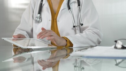 Female doctor, wearing yellow blouse under a medical coat, is using digital tablet in modern healthcare setting, interacting with patient data, close-up. Medicine concept