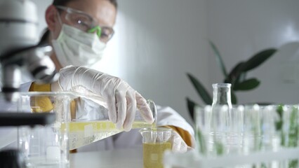 Woman scientist wearing a lab coat, white gloves, and a mask is pouring a yellow oily liquid from one beaker to another in a laboratory setting, close up. Science and medicine