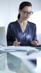 Professional woman accountant in glasses using a calculator and taking notes at a glass desk in the office. Audit and taxes in business