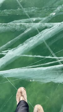 Vertical video of natural ice texture of frozen Baikal Lake on cold winter day. Woman tourist in warm fur boots slowly walks on transparent ice with cracks, sand and algae are visible on bottom