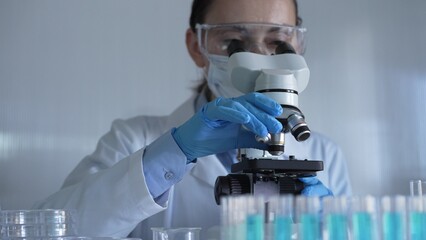 Scientist woman, wearing a lab coat, mask, blue gloves and safety glasses, is adjusting a microscope surrounded by test tubes in laboratory. Medicine and science concept