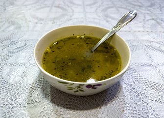 A warm bowl of homemade herbal soup sits on a classic lace tablecloth, evoking the nostalgic charm of a grandmother kitchen. The golden broth, infused with fresh herbs, reflects cozy home cooking