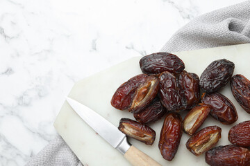 Many tasty dried dates, cutting board and knife on white marble table, top view. Space for text