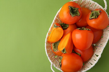 Ripe persimmons in wicker basket on green background, top view. Space for text