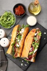 Halves of baguette sandwich with prosciutto and sun dried tomatoes and ingredients on grey table,...