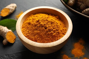 Aromatic turmeric powder in bowl and fresh roots on black table, closeup