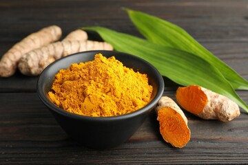 Aromatic turmeric powder in bowl, fresh roots and leaves on wooden table, closeup