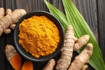 Aromatic turmeric powder in bowl, fresh roots and leaves on wooden table, flat lay