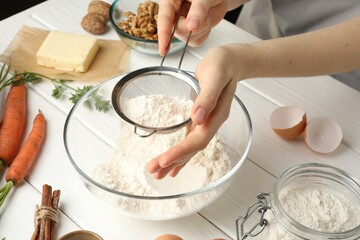 Making carrot cake. Woman sifting flour into bowl at white wooden table, closeup