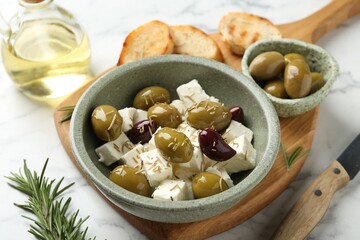 Marinated olives with feta cheese, bread pieces, oil and rosemary on white marble table, closeup