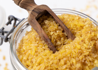 Glass jar of raw dry uncooked yellow bulgur grain with a wooden scoop on napkin closeup