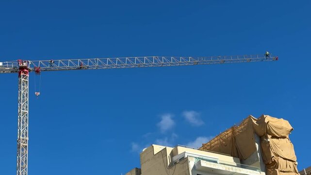 Construction crane against a clear blue sky. On the crane boom there are two workers performing tasks at high heights. The installers are wearing protective equipment, helmets and safety harnesses.