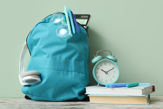 Blue school backpack with different stationery and alarm clock on wooden table near pale green wall