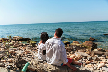 A teenage boy and his mother sitting close on a rocky beach, with his arm around her shoulders, enjoying the picturesque view of the sea under a sunny, cloudless sky