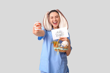 Happy female dentist in bunny ears with Easter basket, greeting card and jaw model on grey background