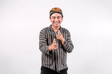A cheerful Indonesian man dressed in traditional Javanese clothing, wearing a striped lurik sorjan and a batik-patterned blangkon, pointing playfully with both hands while smiling in white background.