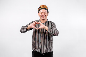 A young man wearing traditional lurik attire smiles brightly while forming a heart shape with his hands, showcasing cultural pride and positivity against a neutral background.