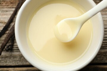 Taking tasty vanilla condensed milk with spoon from bowl on wooden table, flat lay