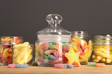 Tasty gummy candies in glass jars on wooden table against grey background, closeup