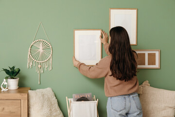 Young woman hanging blank frame on green wall in living room. Back view