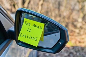 Sticky note with 'The Road Is Calling' on car side mirror