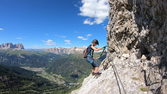 Harnessed to the via ferrata dei finanzieri al Colac, Canazei, Trentino: a climber carefully maneuvers along an exposed ridge, the breathtaking valley far below.