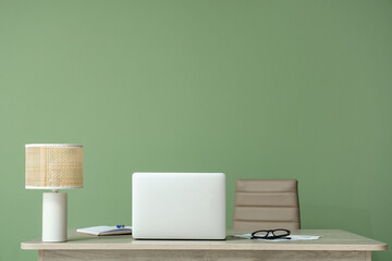 Modern laptop with documents and lamp on table near green wall at home