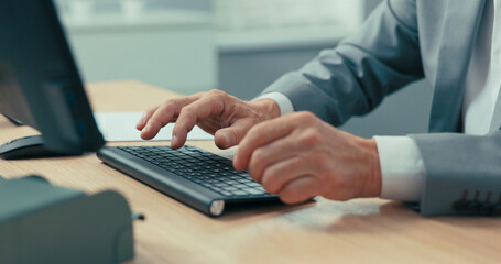 Close-up of older man's hands on black computer keyboard, businessman in gray shirt and jacket,...