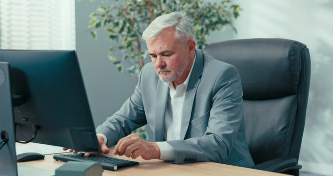 Mature, gray-haired, elegant businessman sits in office at the computer, completes company documents, checks data in the system, sorts invoices, performs managerial duties