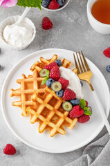Delicious Belgian Waffle with whipped cream, fresh berries and mint on a white plate on a gray concrete background. Summer breakfast.
